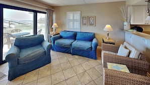 A living room with a blue sofa, a wicker chair, a dining table, and a view through a large window. - Ocean Breeze West #106 | Gulf-Front, Ground Floor, Pool, Beach Access (Perdido Key)