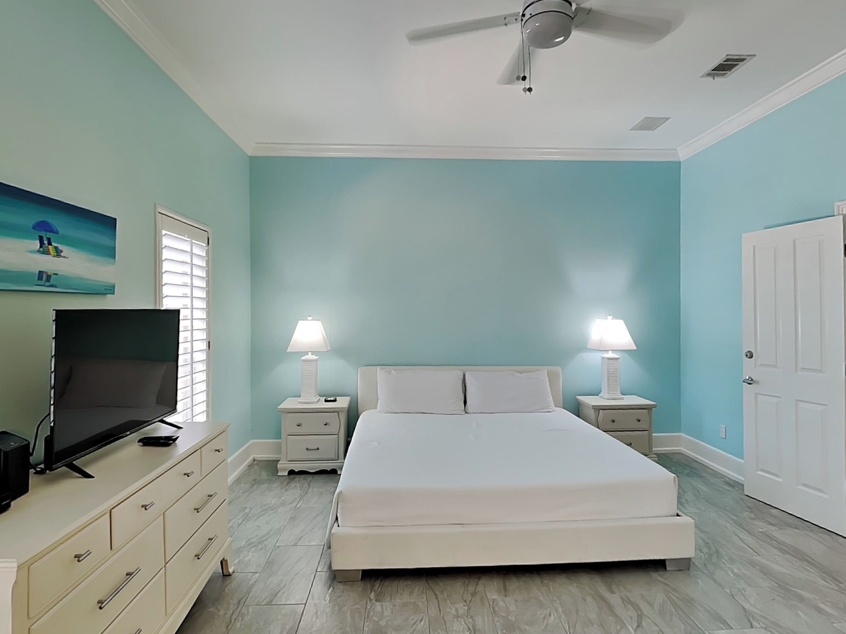 A bedroom with a bed, two bedside tables with lamps, a ceiling fan, a TV on a dresser, and a door.