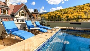 Un área de piscina con sillas reclinables azules, una piscina con azulejos y montañas al fondo.