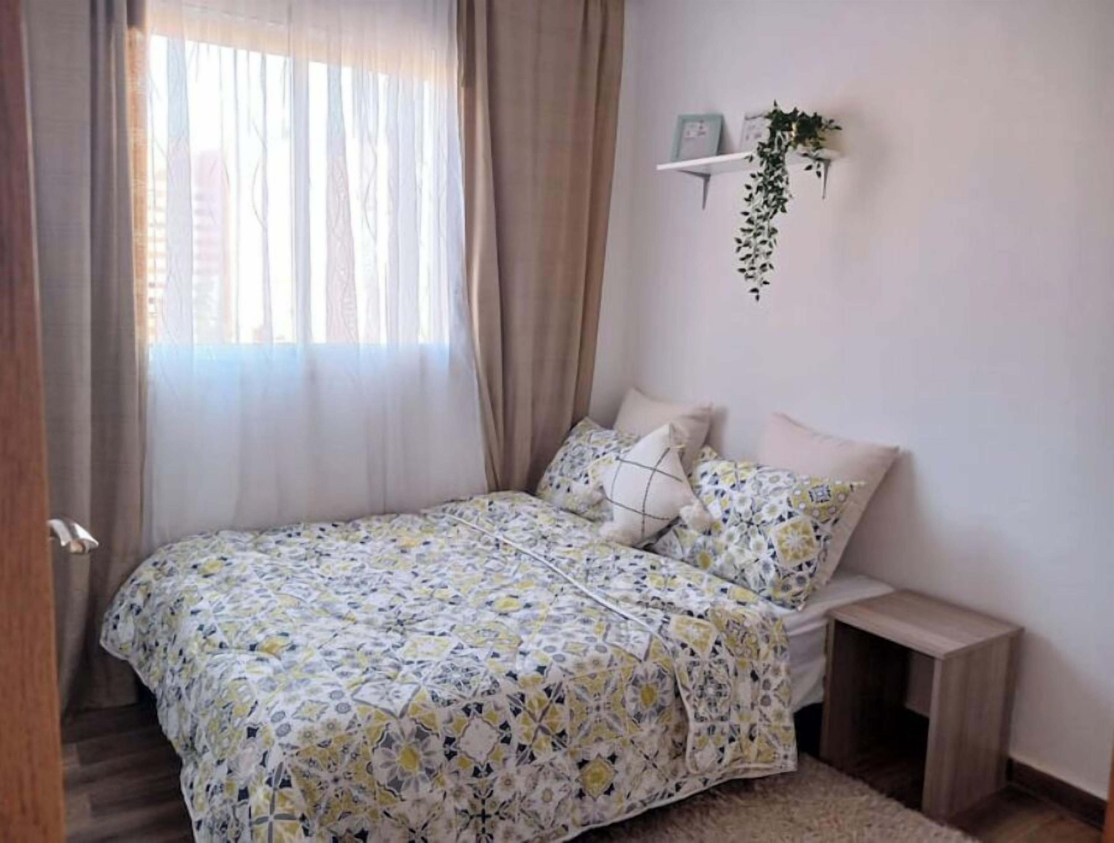 A bedroom with a bed, a bedside table, a hanging plant, and a window with curtains.