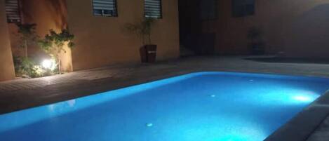 A swimming pool at night with a blue illuminated edge, surrounded by a building with windows and potted plants.