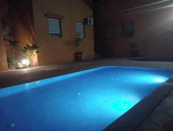 A swimming pool at night with a blue illuminated edge, surrounded by a building with windows and potted plants. - Appartement de Charme (Marrakech)