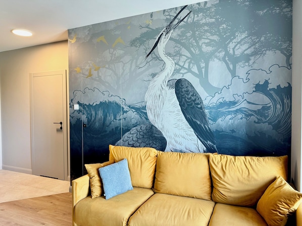 Habitación con un mural grande de un pájaro y árboles, un sofá con cojines y una puerta.
