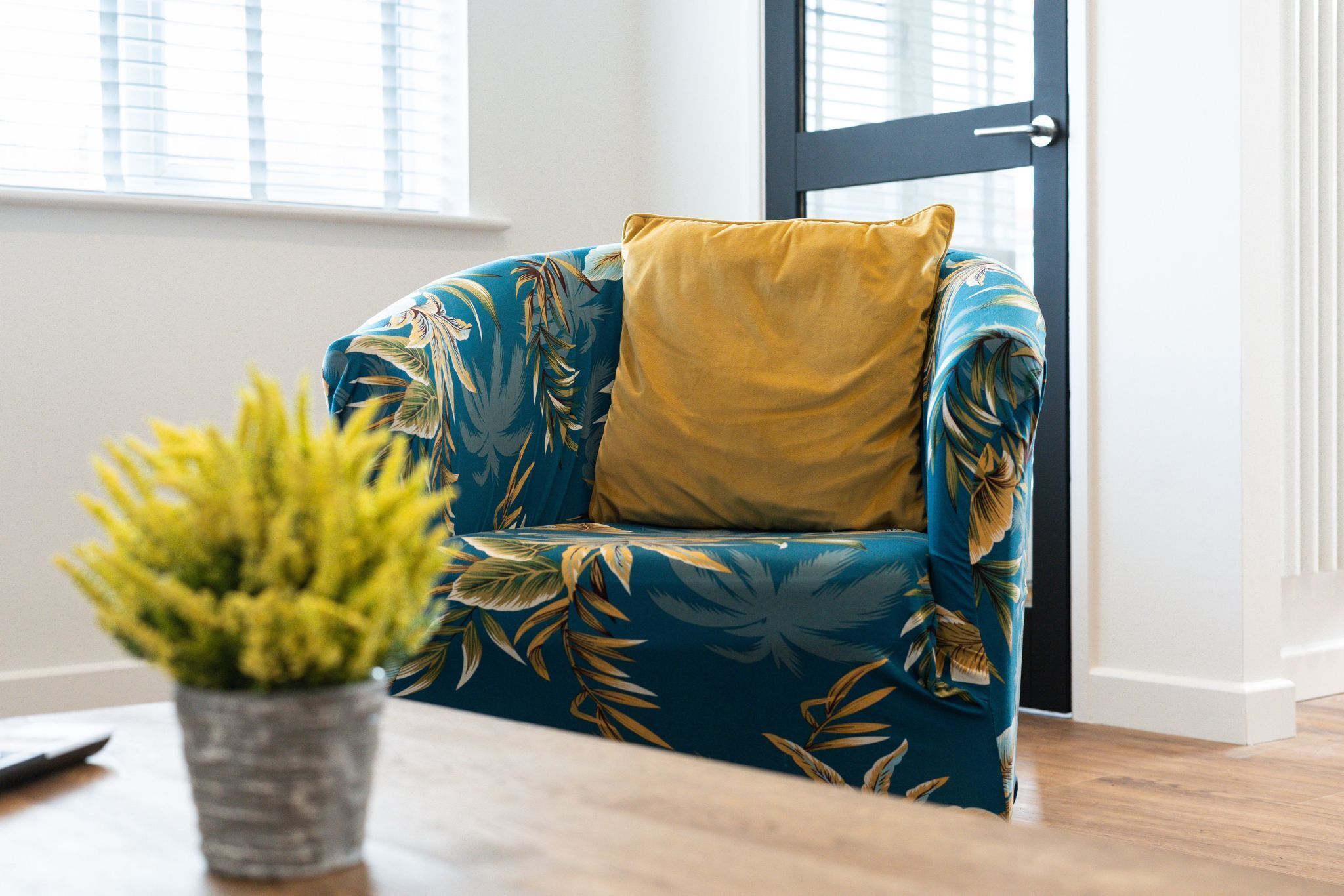 A room with a blue floral sofa, a yellow pillow, and a potted plant on a wooden table.