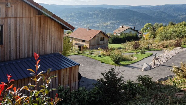 Una casa de madera con techo inclinado, rodeada de un jardín cuidado y vistas a montañas distantes.