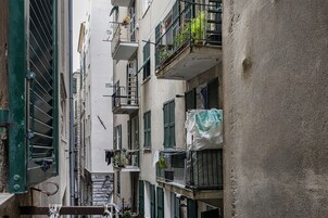 Una estrecha calle urbana flanqueada por edificios de varios pisos, con balcones adornados con plantas y ropa tendida.