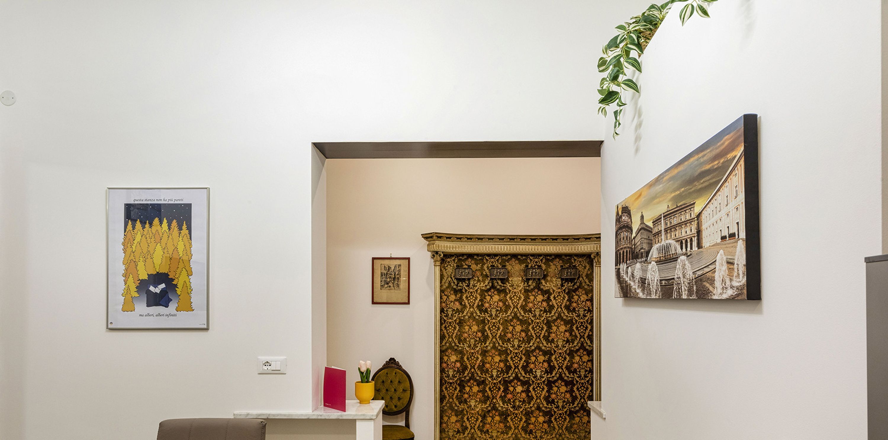 A room with a white wall, a framed picture of a forest, a framed picture of a building, a plant hanging from the ceiling, and a chair with a cushion.