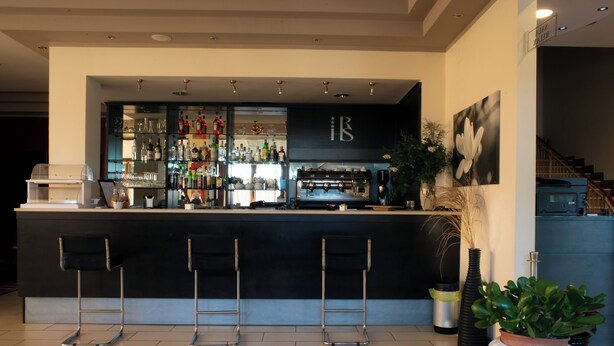 A modern bar area with a black countertop, three bar stools, a coffee machine, and a variety of bottles and glasses. - HOTEL HRS (Follonica)