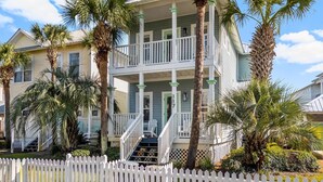 A two-story house with a balcony, surrounded by palm trees and a white picket fence.
