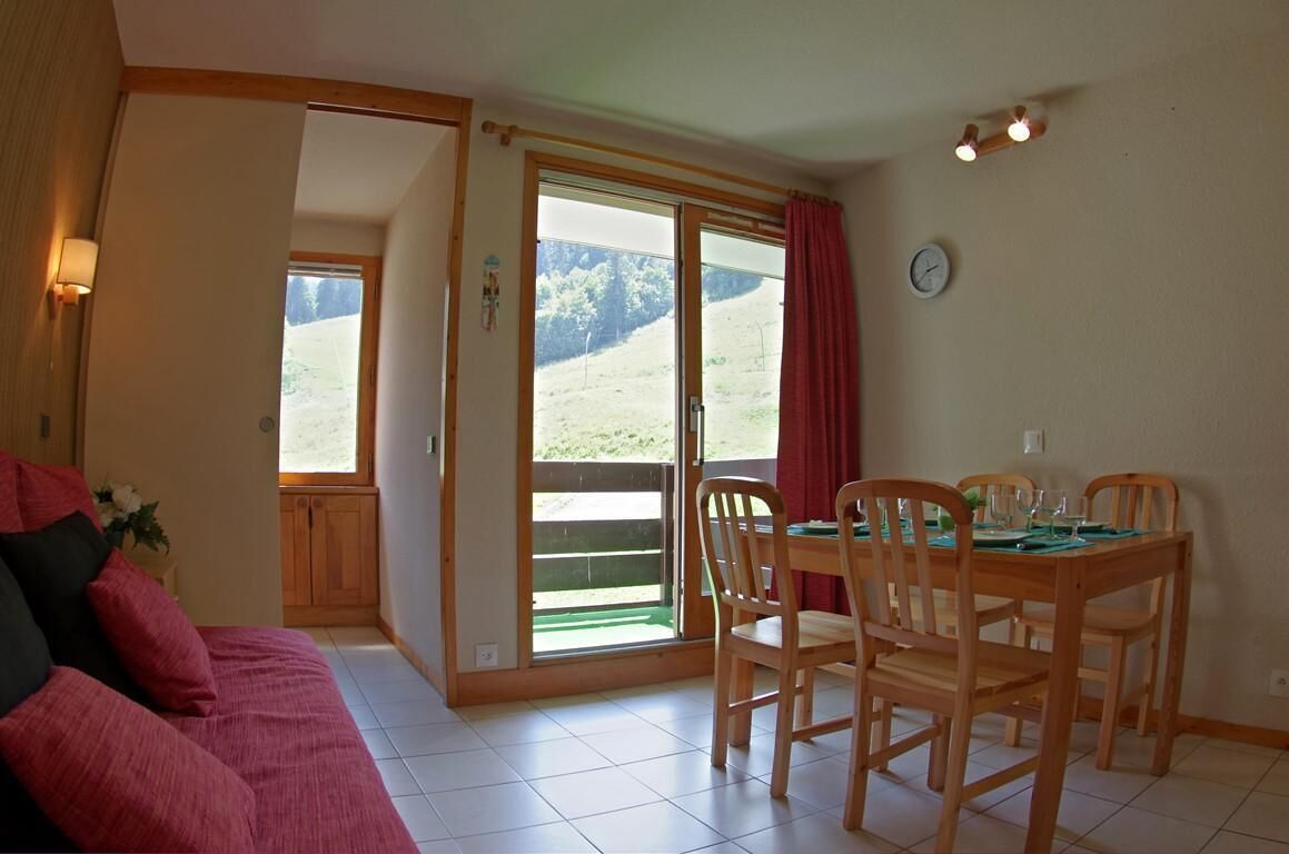 Une pièce chaleureuse avec une table à manger en bois, préparée pour quatre personnes, un canapé rouge et une grande porte-fenêtre coulissante donnant sur un balcon avec vue sur un paysage verdoyant.