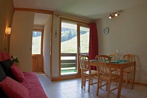 A cozy room with a wooden dining table set for four, a red sofa, and a large sliding glass door leading to a balcony with a view of a green landscape. - 1st floor, view ski slopes, balcony, tv, 25m², Valmorel (Valmorel)