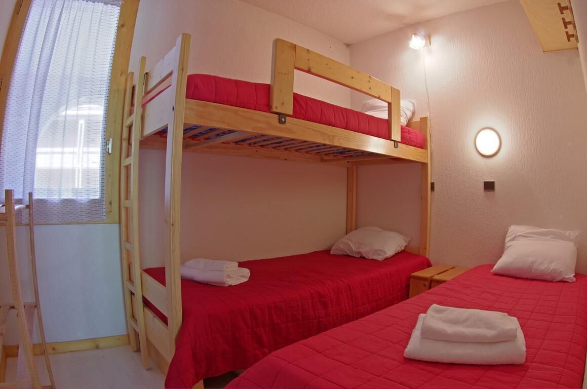Une chambre avec deux lits superposés, chacun recouvert d’une couverture rouge et agrémenté d’oreillers blancs. On y trouve une petite table de chevet en bois sur laquelle est posée une lampe.