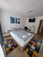 Un dormitorio moderno con una cama de madera, paredes blancas, un televisor de pantalla plana y una alfombra colorida.