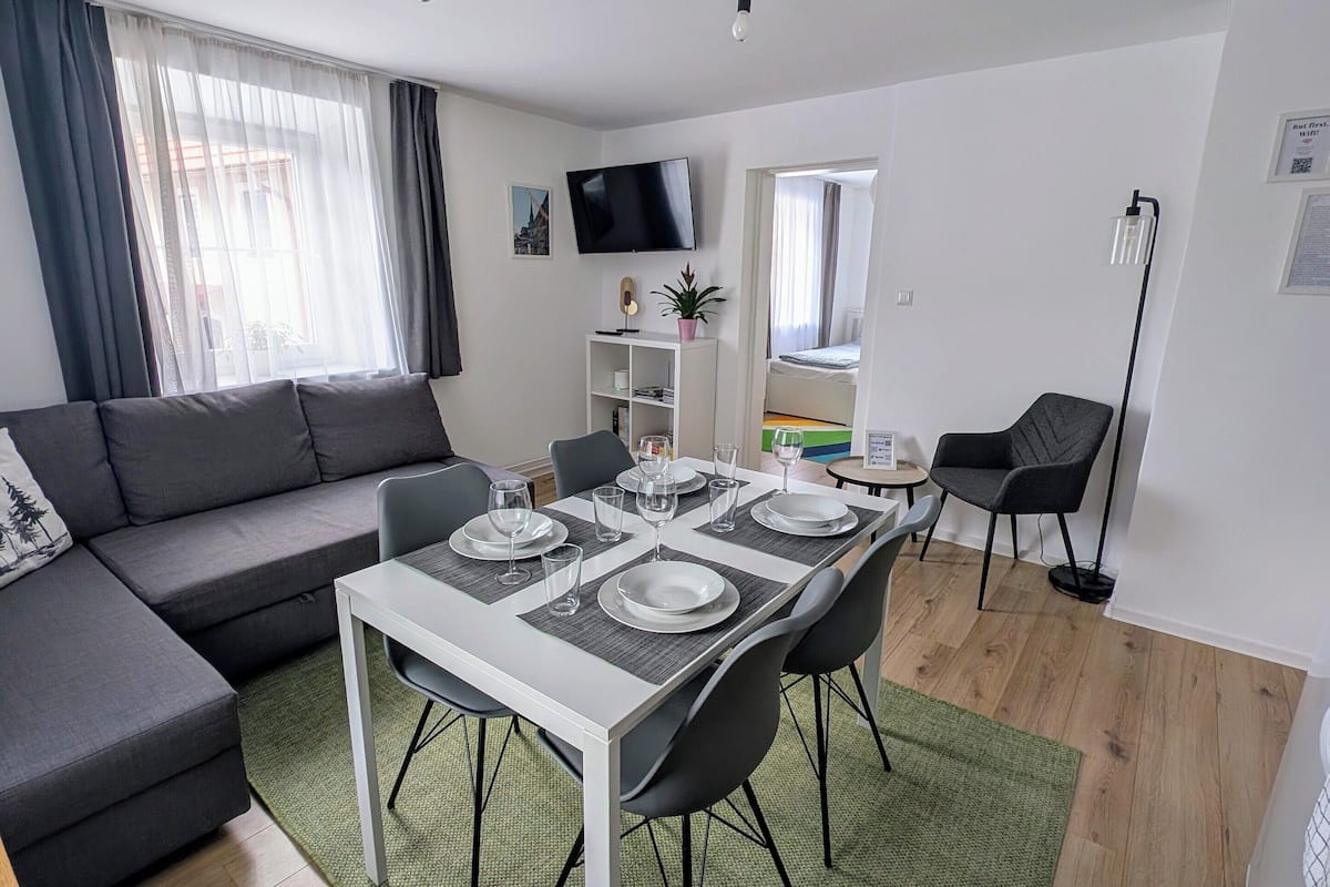 A modern living room with a dining area, grey sofa, flat-screen TV, and a green rug.