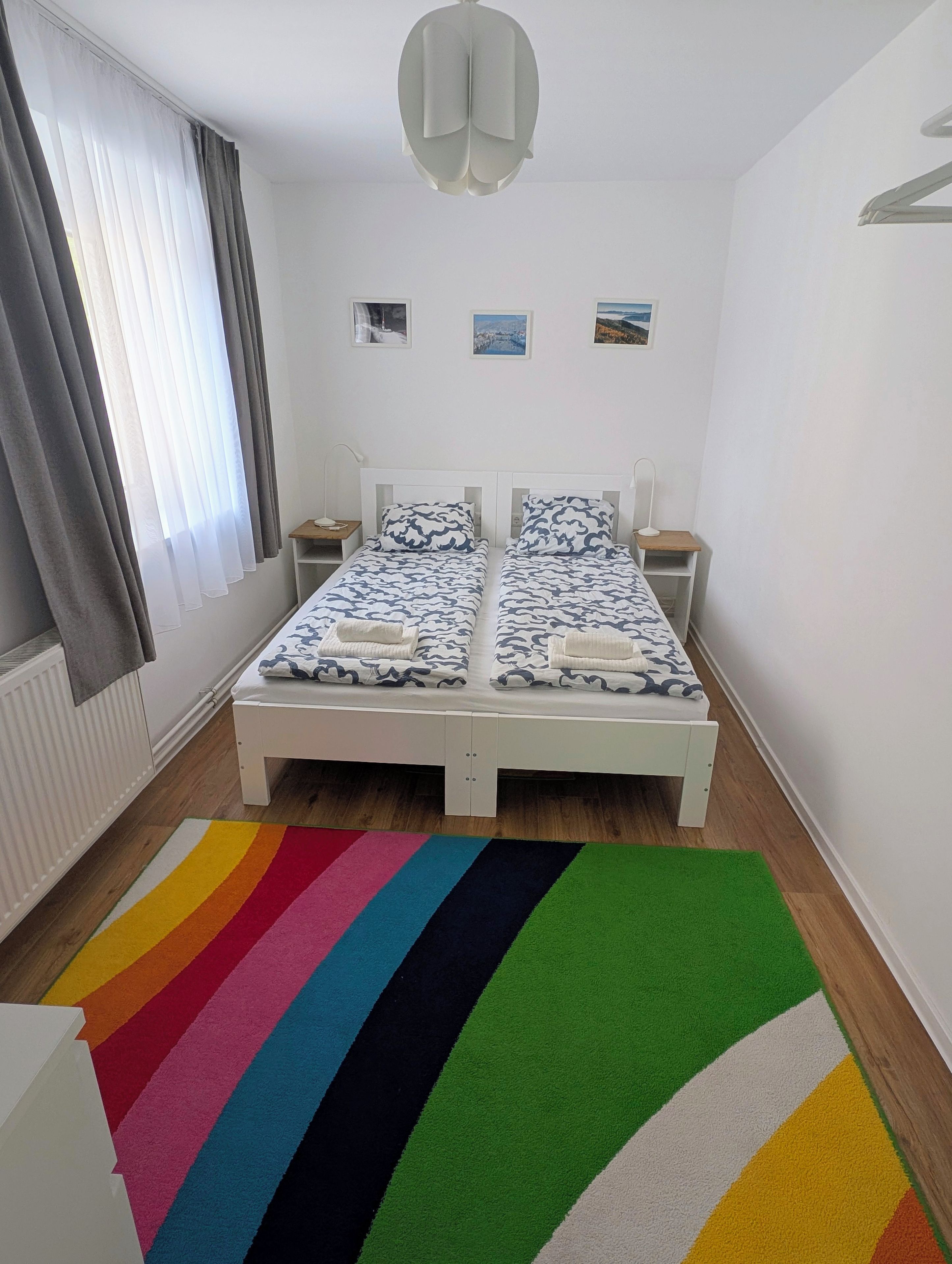 Une chambre à coucher avec un lit, deux tables de chevet, une commode, un radiateur, une fenêtre avec des rideaux et un tapis coloré.