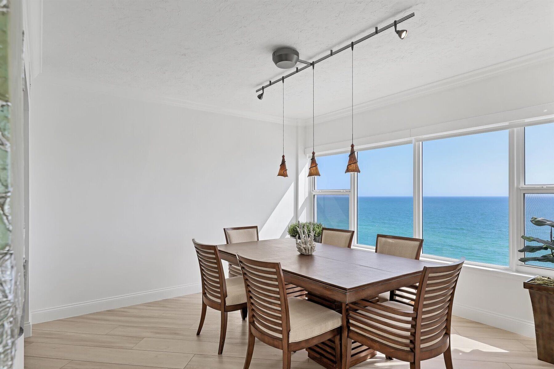 Un espace repas avec une table et des chaises en bois, de grandes fenêtres donnant sur l  ’océan et des luminaires suspendus.