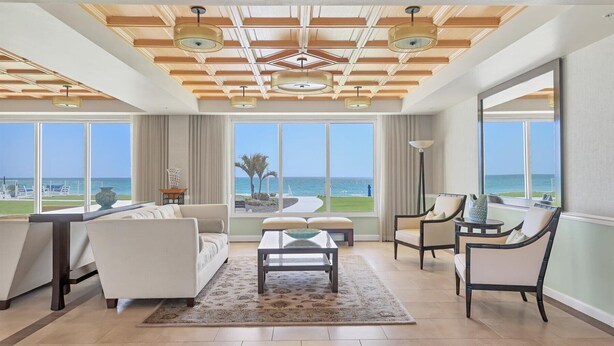 Un salón amplio con un gran ventanal con vista al océano, techo con vigas de madera y lámparas colgantes, y una distribución moderna de los muebles.