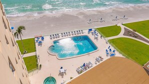 Vista aérea de un resort con piscina central rodeada de sillas de descanso y sombrillas, zona de playa y un edificio con balcones.
