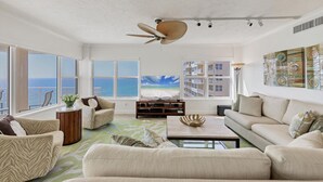 Un salón amplio con un gran ventanal con vista al océano, un ventilador de techo y un televisor de pantalla plana.