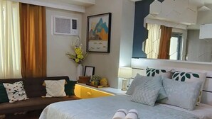 Habitación de hotel con cama, mesita de noche, una silla, lámpara en el techo, ventana con cortinas, aire acondicionado instalado en la pared y un cuadro enmarcado.
