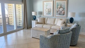 Un salon spacieux avec un mur bleu clair, un canapé beige, deux fauteuils, une table basse et deux petites tables d’appoint avec des lampes. Deux tableaux encadrés ornent le mur, et un ventilateur de plafond est visible.