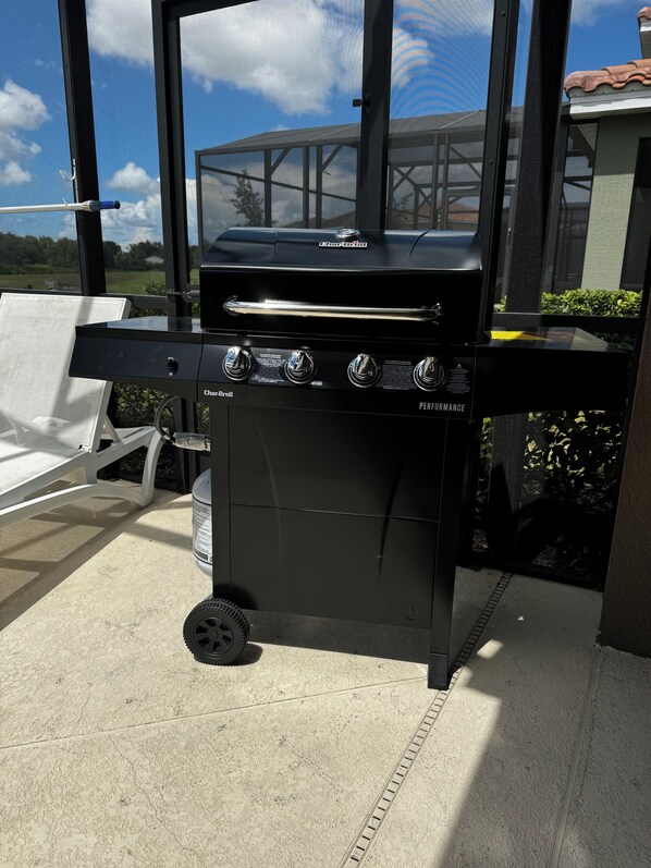 Una parrilla de gas Char-Broil con cuatro quemadores, ubicada en un patio que ofrece vistas a una casa y follaje.