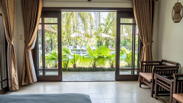 A room with a bed, a dining table with chairs, and a view of a garden with palm trees and umbrellas. - Riverside Bamboo Resort - Hoi An (Da Nang)
