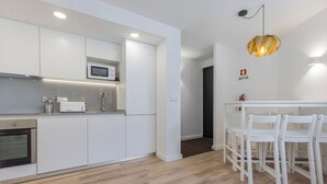 Una cocina moderna con armarios blancos, microondas y horno empotrado. Hay una zona de comedor con sillas blancas y una mesa.
