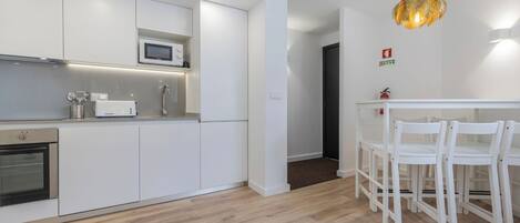 Una cocina moderna con armarios blancos, microondas y horno empotrado. Hay una zona de comedor con sillas blancas y una mesa.