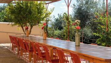 An outdoor dining area with a long wooden table and red chairs, surrounded by greenery and trees.