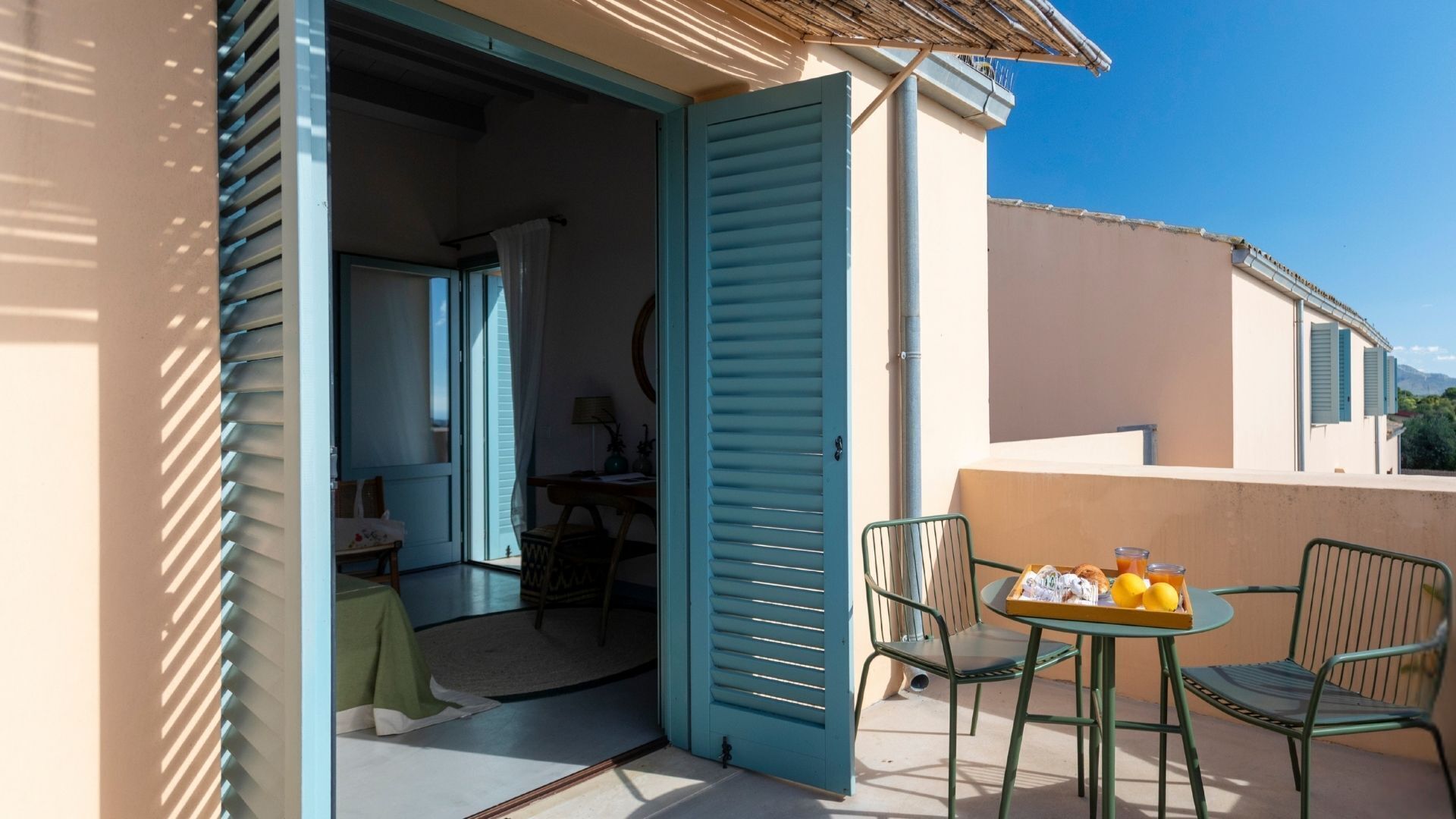 An open door leading to a room with a table and chairs, a balcony with a table set for breakfast, and a view of the surrounding area.