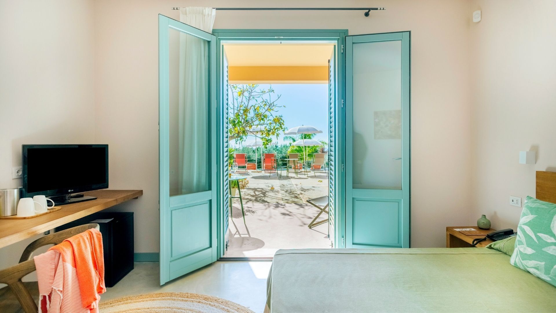 A hotel room with a bed, a desk with a TV, and a view of an outdoor seating area with umbrellas.