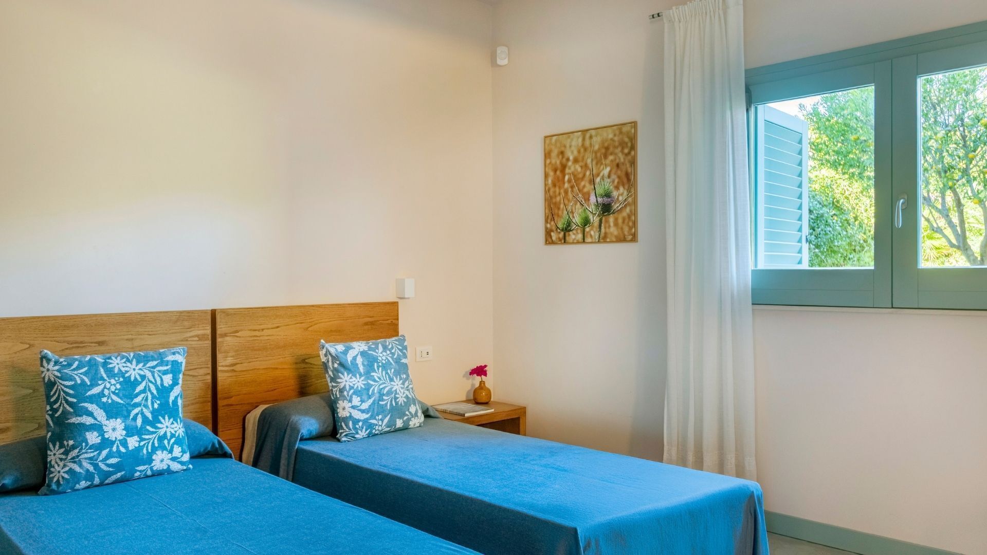 A room with two beds, a wooden headboard, a nightstand, a vase with flowers, a painting, and a window with curtains.
