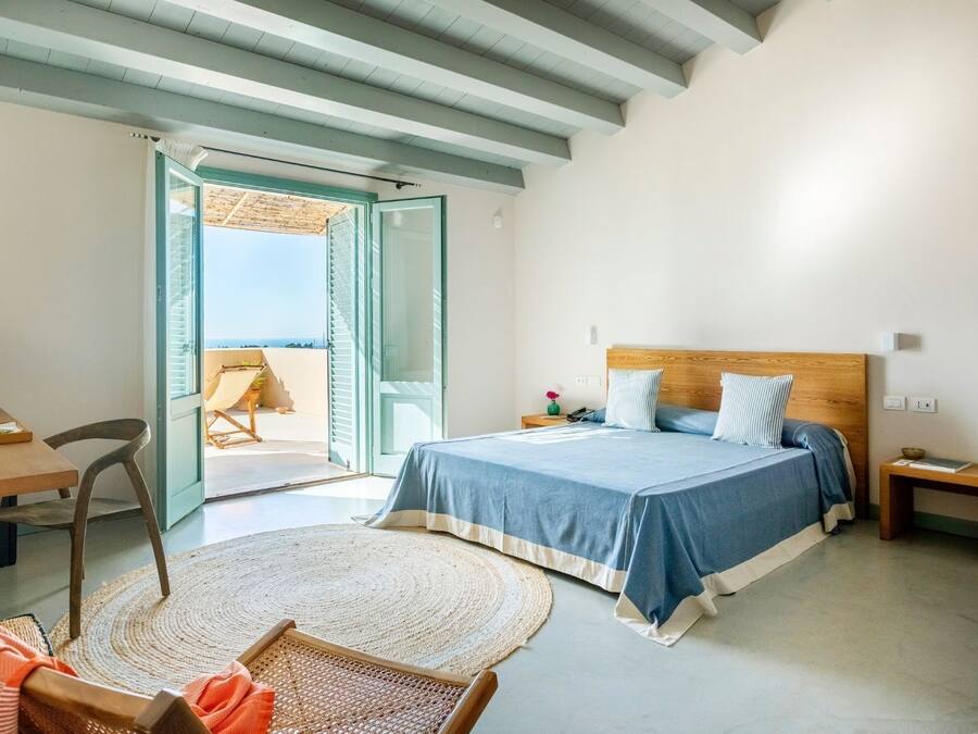 A bedroom with a bed, a desk, a chair, a small table, and a view of the outside.