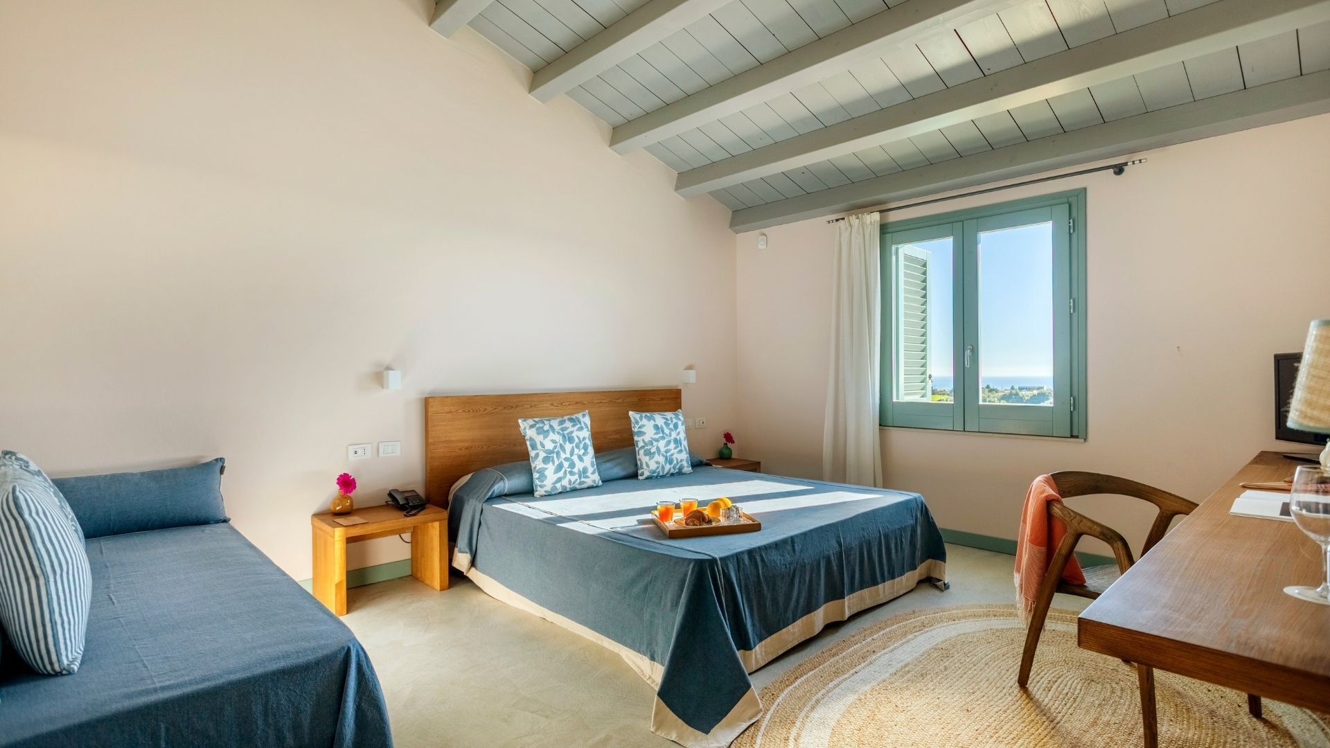Un dormitorio con una cama, una mesita de noche, una silla, una pequeña mesa con un vaso y vista al mar.