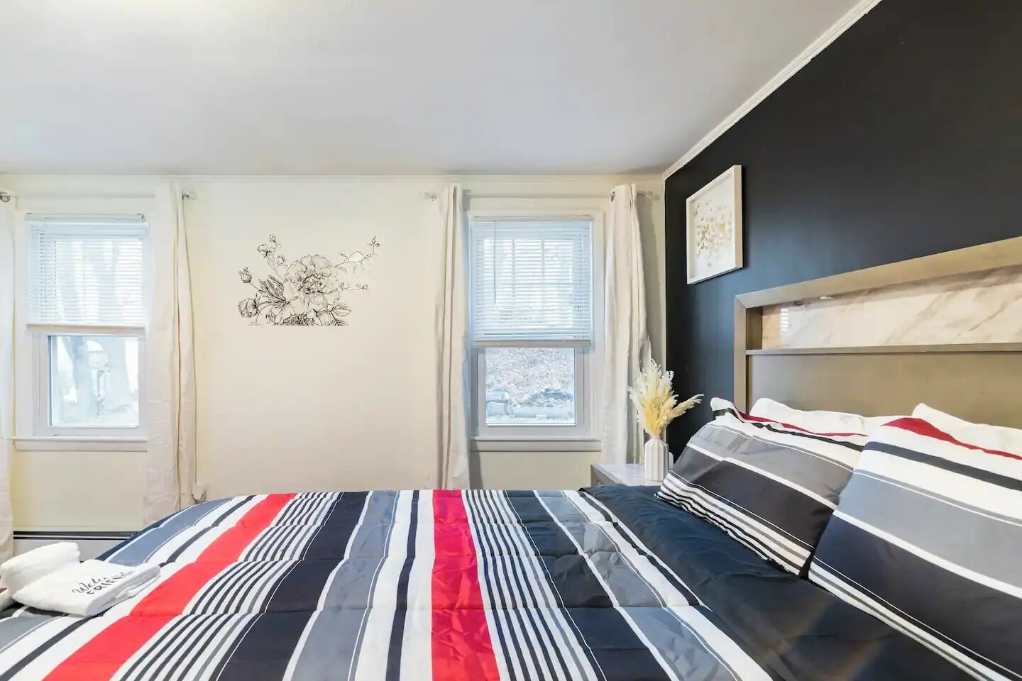 Une chambre à coucher avec un lit, deux fenêtres, un élément de décoration murale et une table de chevet sur laquelle se trouve un vase.