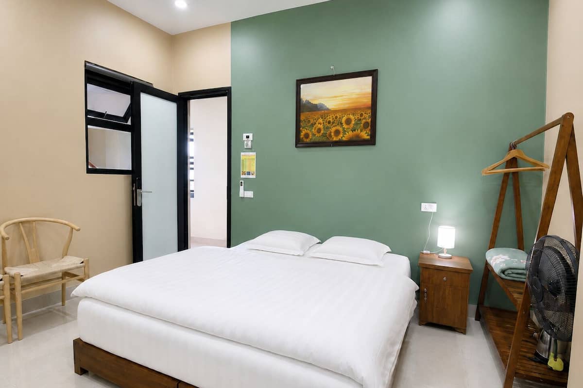 A bedroom with a bed, a chair, a wooden hanger, a fan, and a painting on the wall.