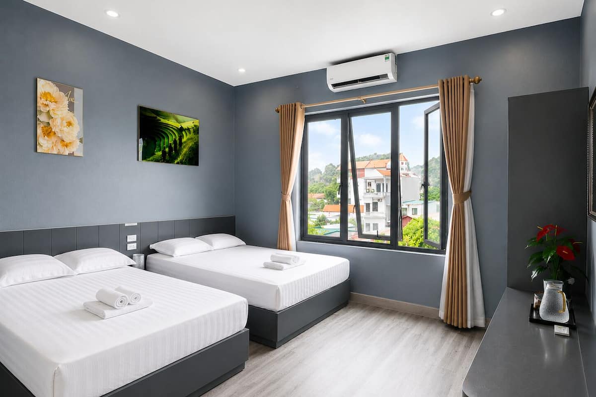 A hotel room with two beds, a large window with curtains, an air conditioning unit, and a desk with a vase of flowers.