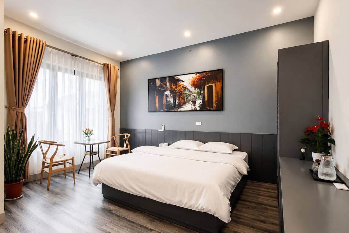 A modern hotel room with a large bed, a dining table, chairs, a potted plant, and a painting on the wall.