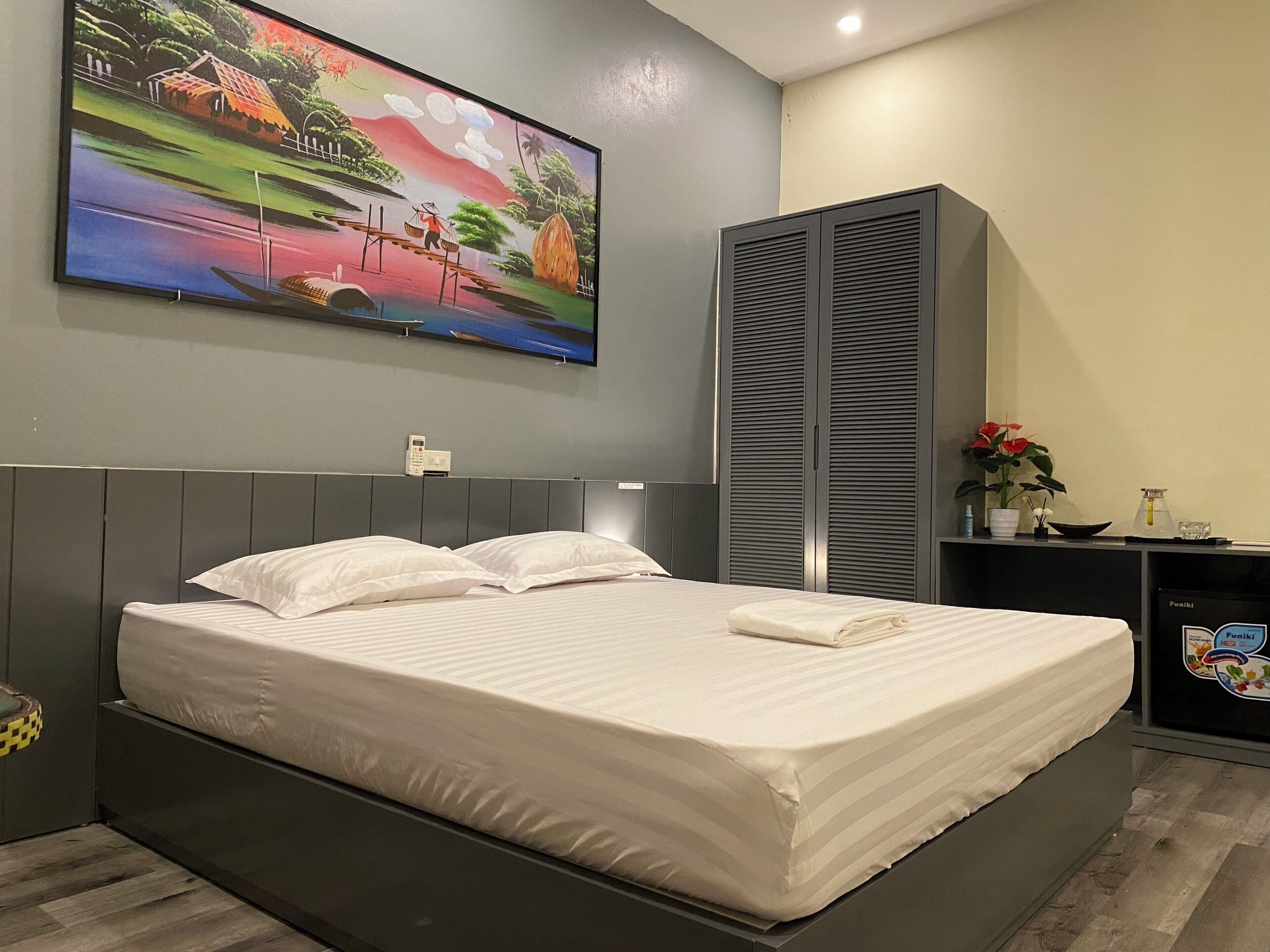 A modern hotel room with a large bed, a flat-screen TV mounted on the wall, a bedside telephone, and a small desk with a plant and a water bottle.