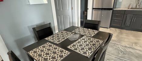 Un coin repas avec une table dressée pour quatre personnes, entourée de chaises. On peut voir un panneau indiquant la présence de matériel de premiers secours sur le mur, ainsi qu’un réfrigérateur et un four à micro-ondes dans la cuisine.