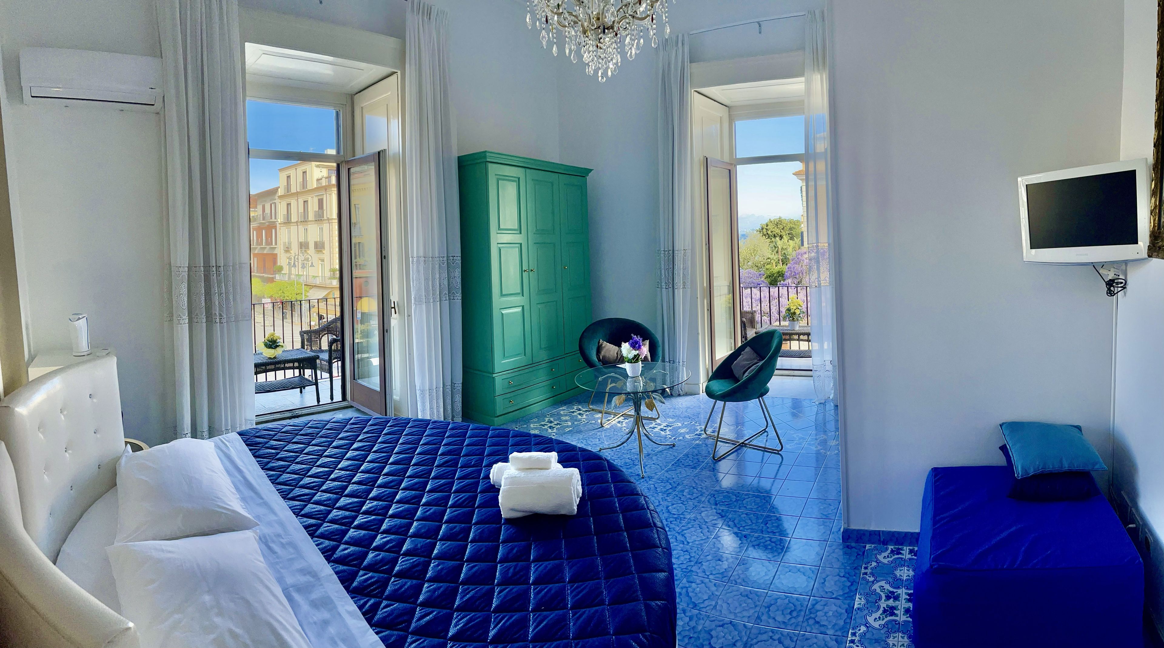 Una camera d'hotel con un letto, un'armadio verde, un tavolino in vetro con una sedia e una TV a parete.