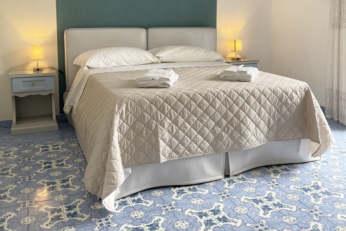 A neatly made bed with a quilted cover, two bedside lamps, and a wall-mounted air conditioner.