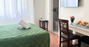 Une chambre d’hôtel avec un lit recouvert d’une courtepointe verte, une chaise en bois, une petite table avec des articles pour le petit-déjeuner et une fenêtre ornée de rideaux légers.