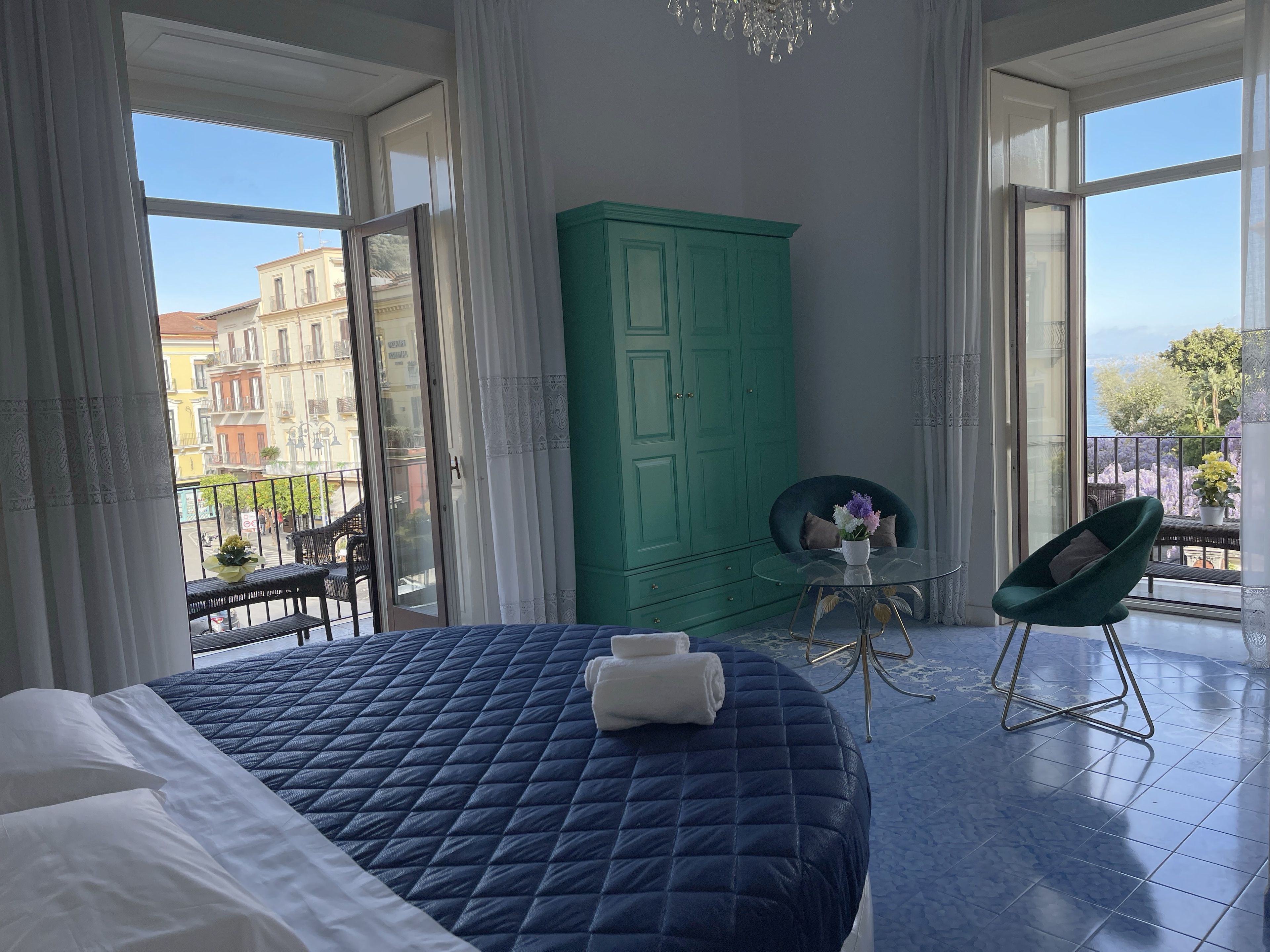 A bedroom with a large bed, a green wardrobe, a small table with a chair, and a balcony with a view of buildings.