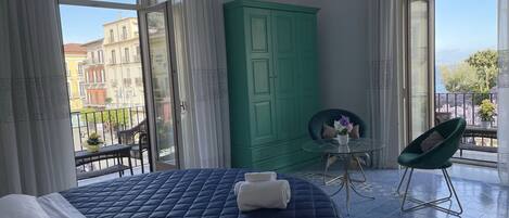 A bedroom with a large bed, a green wardrobe, a small table with a chair, and a balcony with a view of buildings.