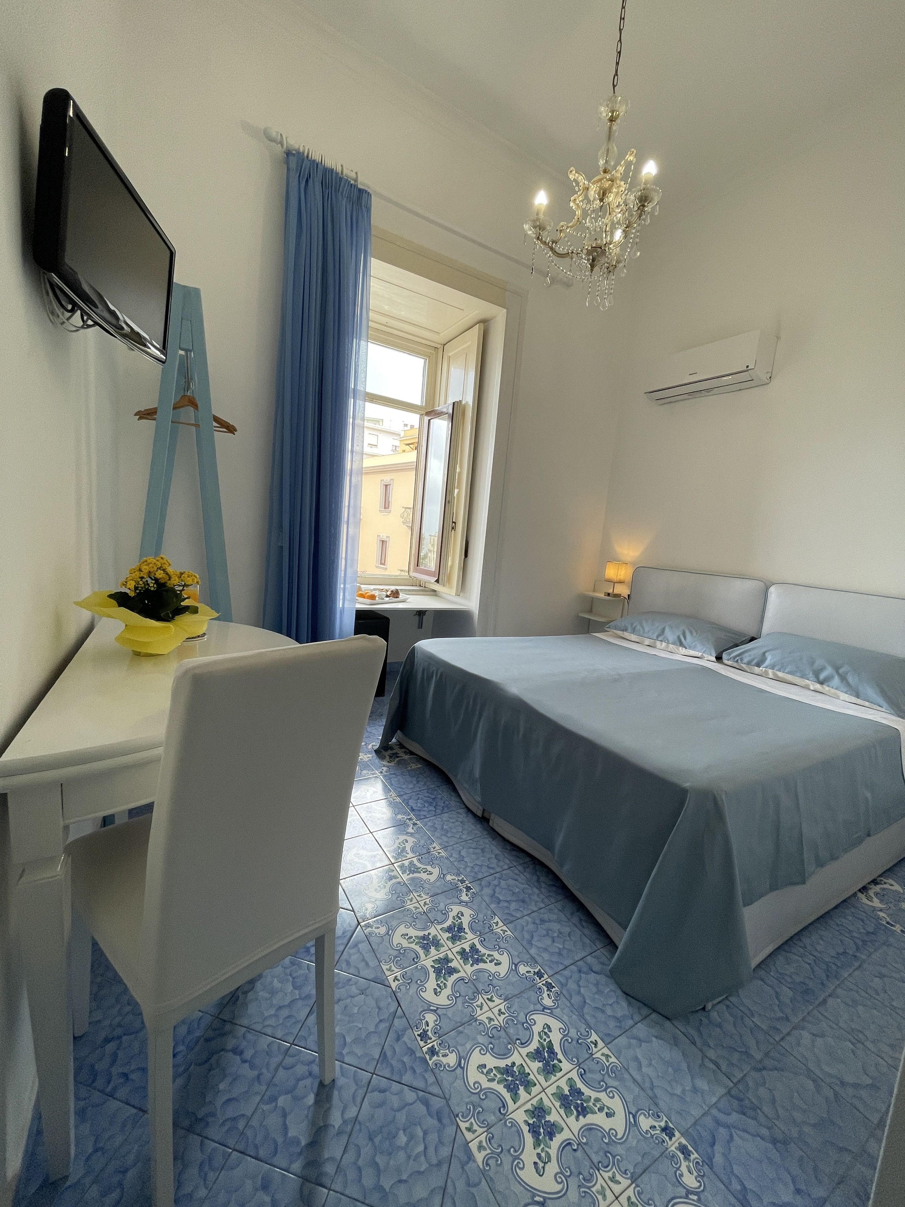 A hotel room with a bed, a desk, a chair, a TV, a window with curtains, and a chandelier.