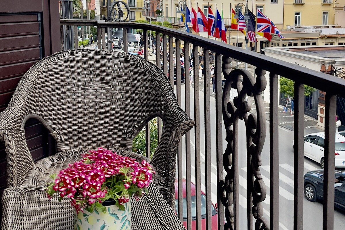 Ein Balkon mit Korbmöbeln und einem Blumentopf, mit Blick auf eine Straße mit Gebäuden und Flaggen.