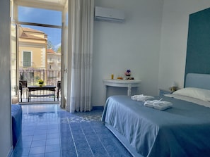 Ein Hotelzimmer mit Bett, Nachttisch mit Lampe, Fenster mit Vorhängen, Balkon mit Stuhl und Tisch sowie Wandgerät zur Klimatisierung.