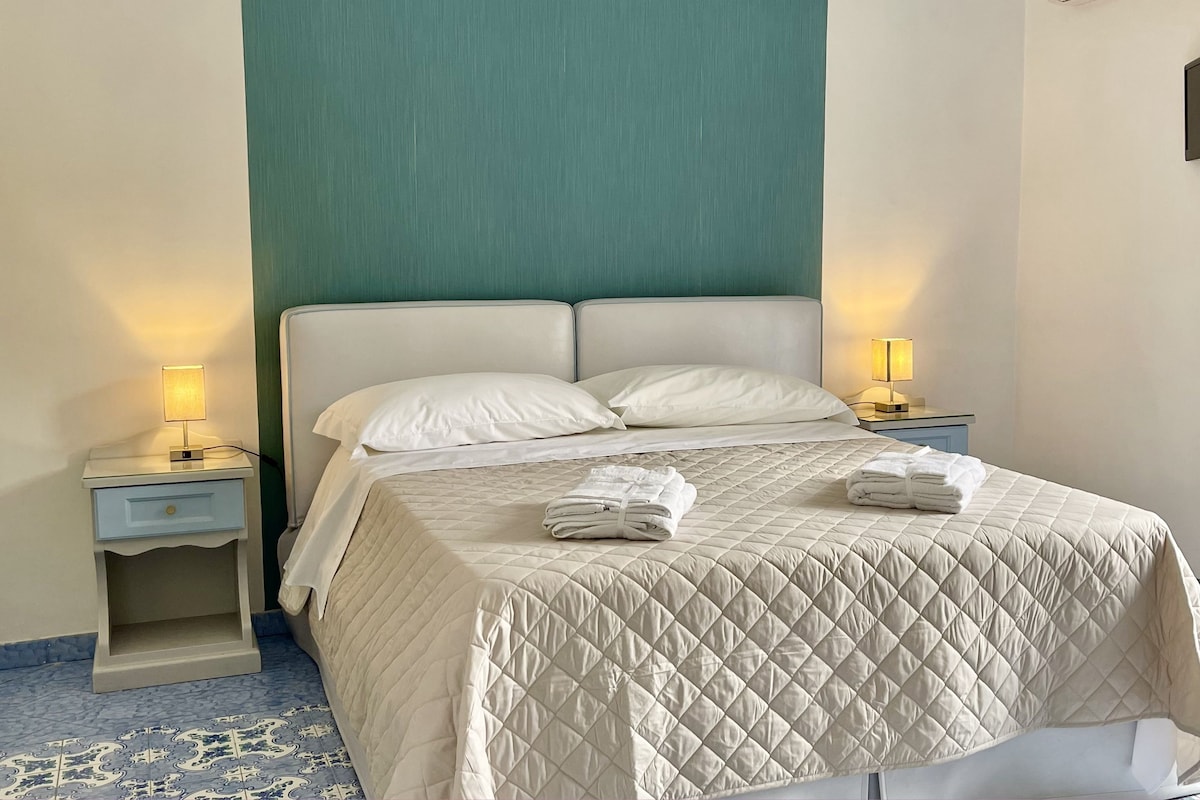Un lit bien fait, recouvert d'une courtepointe matelassée, avec deux oreillers et une pile de serviettes pliées posée sur le pied de lit. Il y a deux tables de chevet avec des lampes, une de chaque côté du lit.